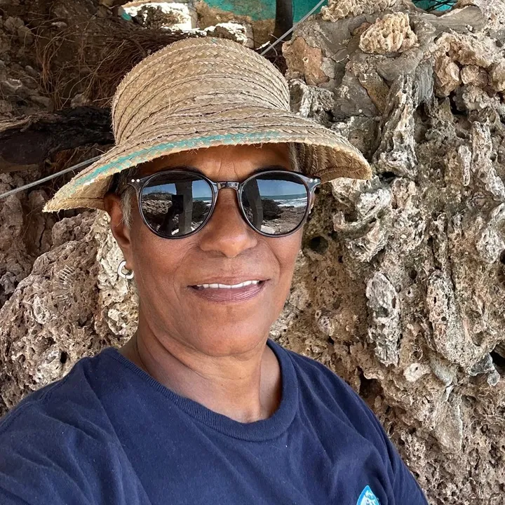Annie Reynolds in a straw visor and sunglasses, in front of a wall of beach rock