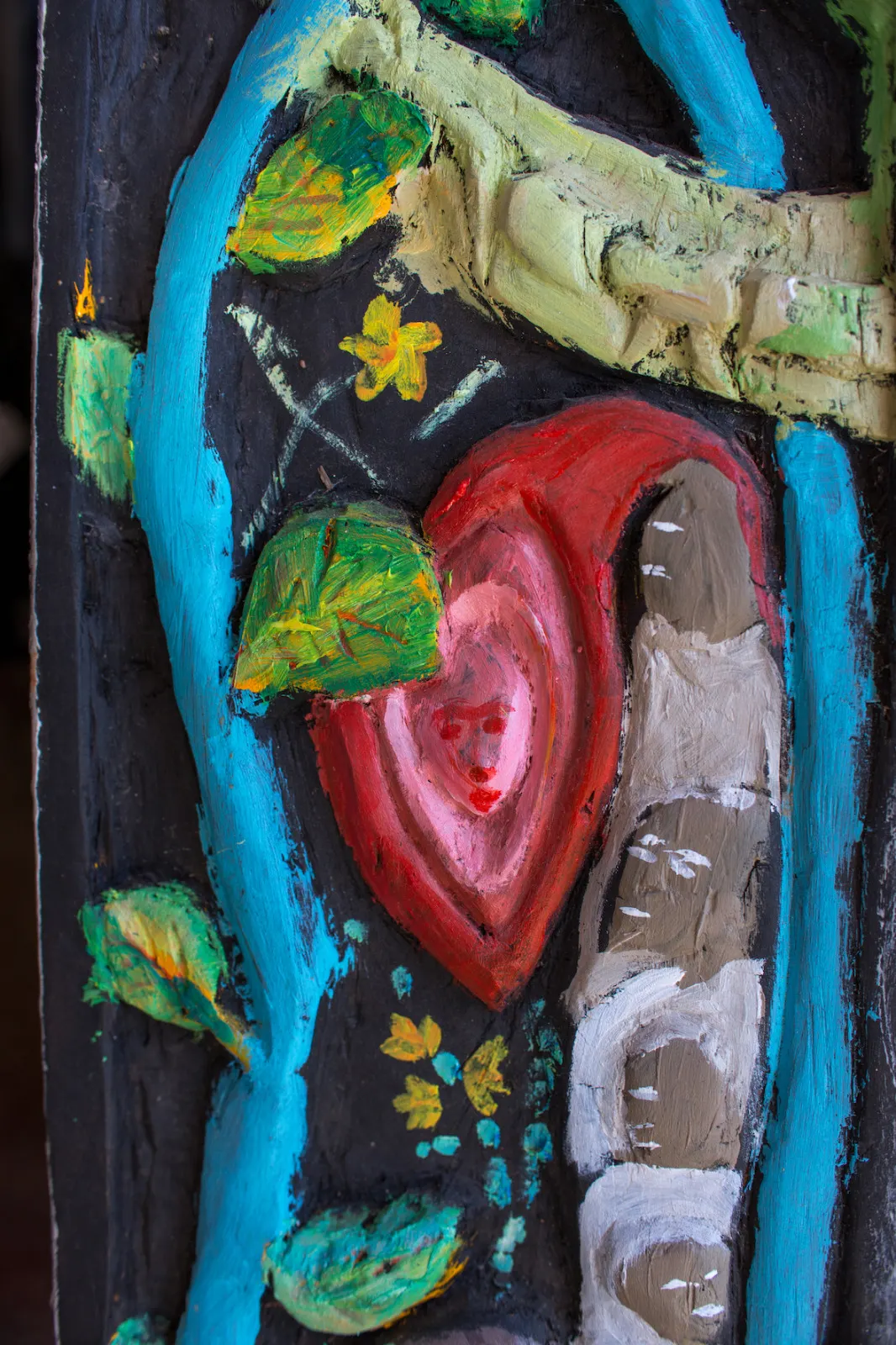 A carved wooden relief painted with a red heart holding a face, blue branches and green leaves