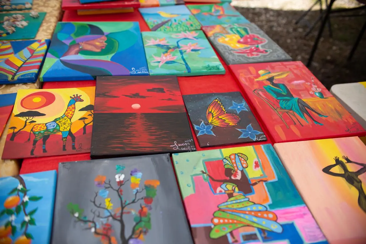A table of his paintings: a giraffe, a red sunset, a butterfly, a woman in a hat, and more, signed Lucas Williams