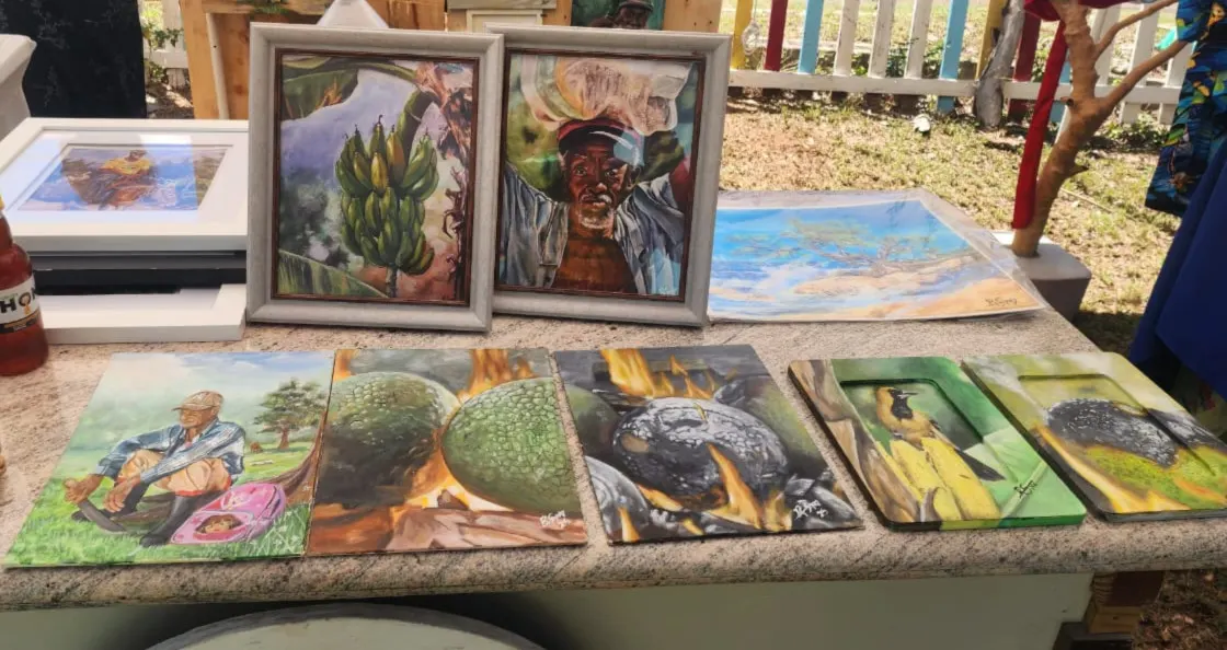 Framed and unframed paintings on a table: bananas, a man carrying a load, breadfruit roasting and a bird