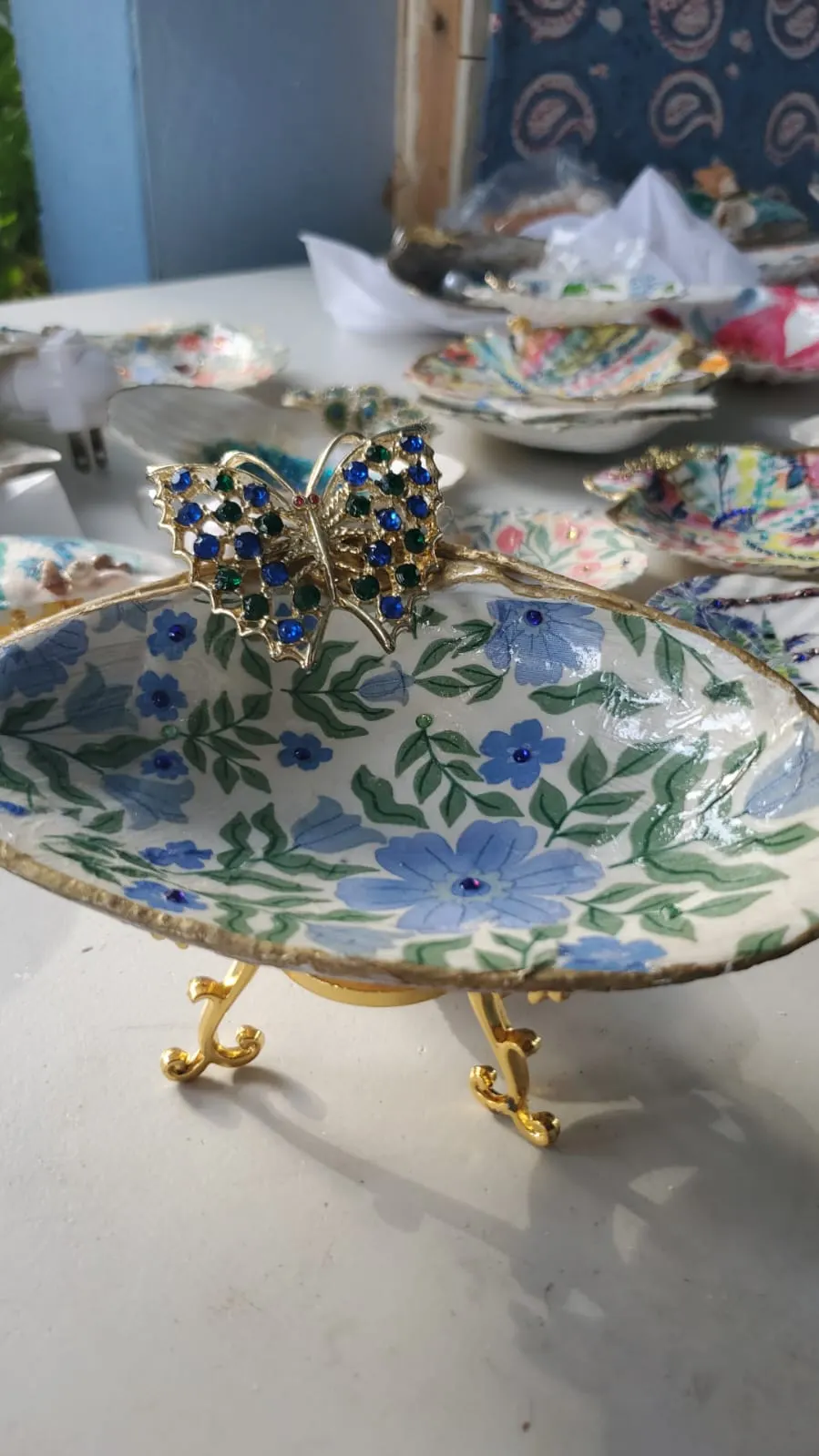 A shell dish on gold feet decorated with blue flowers, with a jewelled butterfly on its rim
