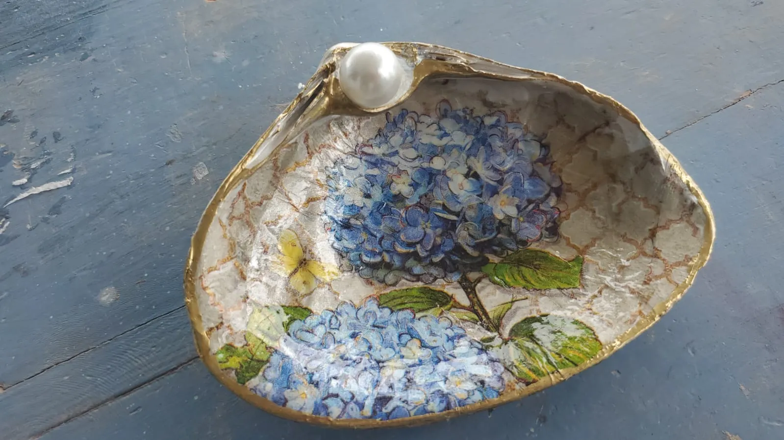 A shell dish decorated with blue hydrangeas, a butterfly and a pearl, edged in gold