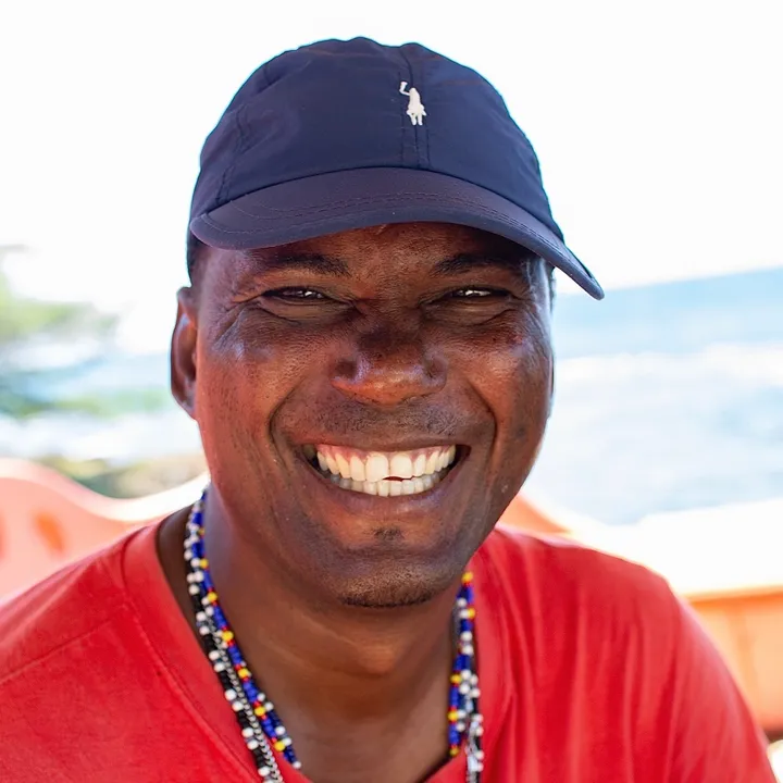 Sandy Straughn, smiling, in a navy cap and red T-shirt, by the sea