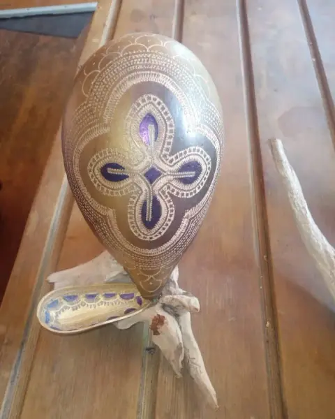 A calabash carved with a fine white pattern in gold and purple, with a matching calabash spoon, on driftwood