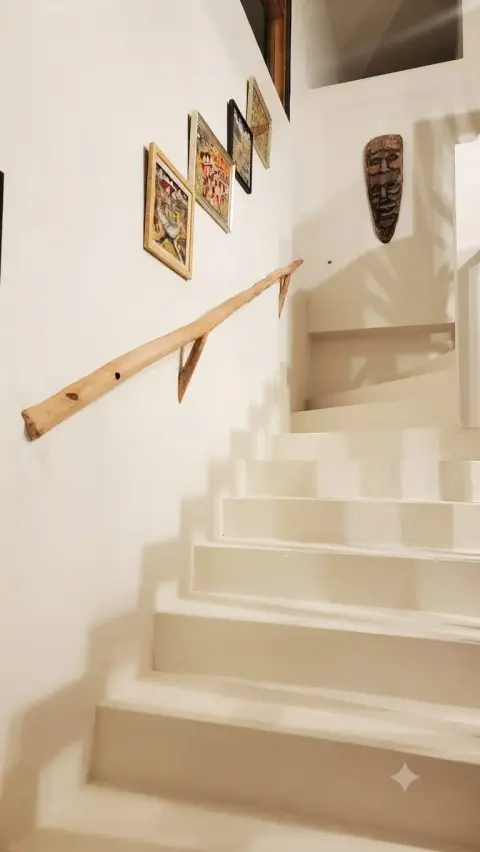 A driftwood handrail along a white staircase, under framed paintings and a carved mask