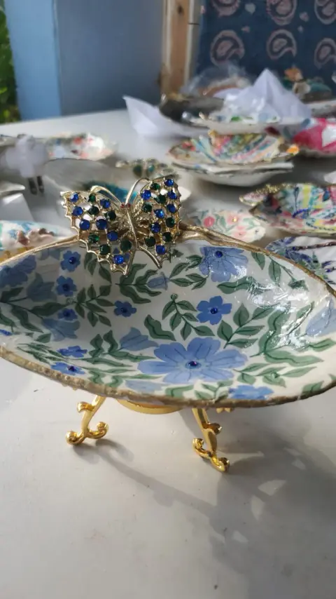 A shell dish on gold feet decorated with blue flowers, with a jewelled butterfly on its rim