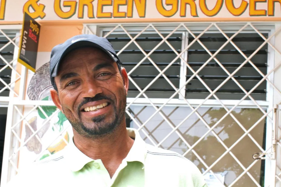 Captain Dennis smiling at the camera, in a dark cap and a light green polo shirt, in front of a shop with a painted sign and a white security grille