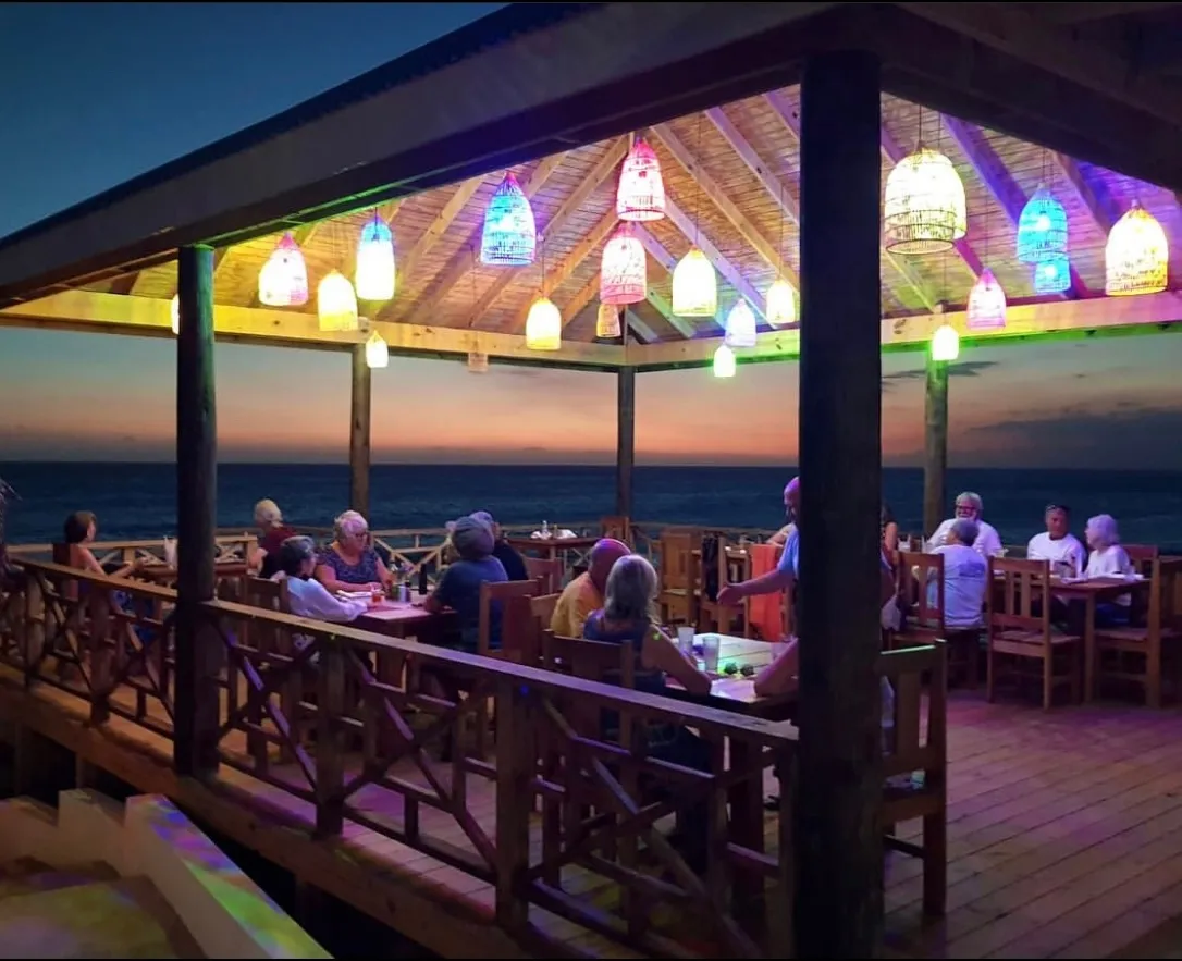 Beyond Sunset Bar and Smoke Grill — The restaurant deck at dusk, with coloured lamps lit and guests at the tables, and the sea behind them