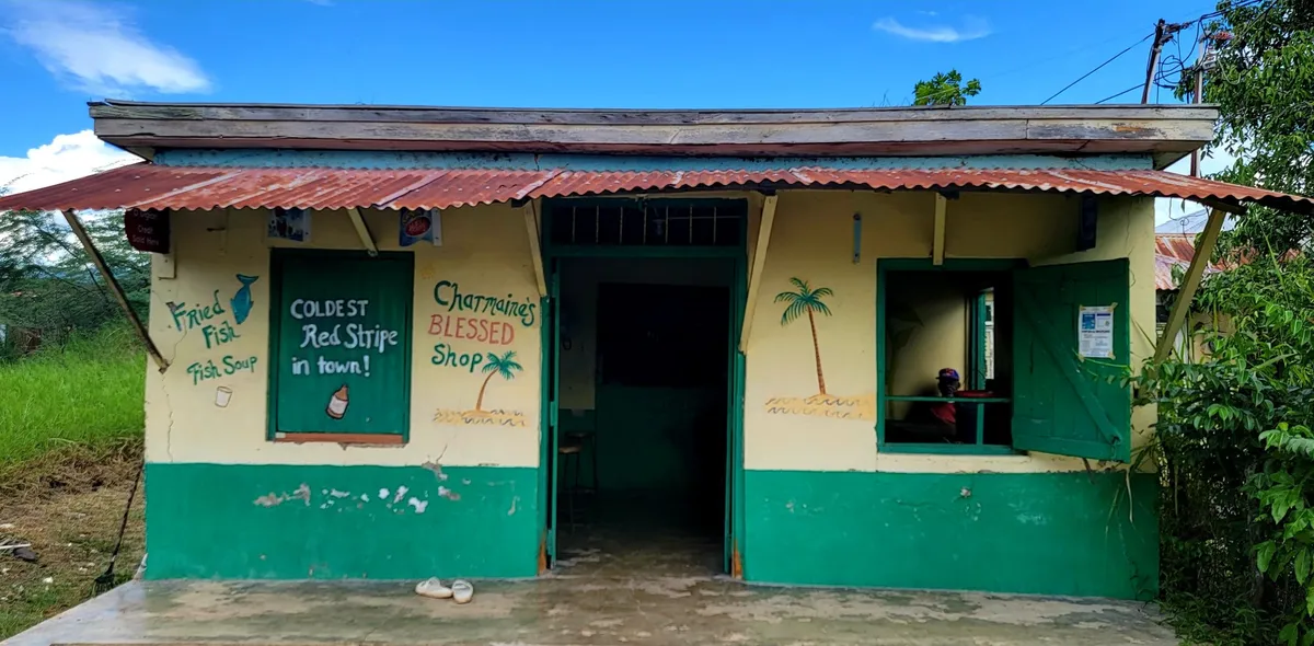 Charmaine’s Blessed Shop, a yellow and green painted building with a rusty zinc awning, hand-painted with Fried Fish, Fish Soup, Coldest Red Stripe in town and the shop name, under a blue sky