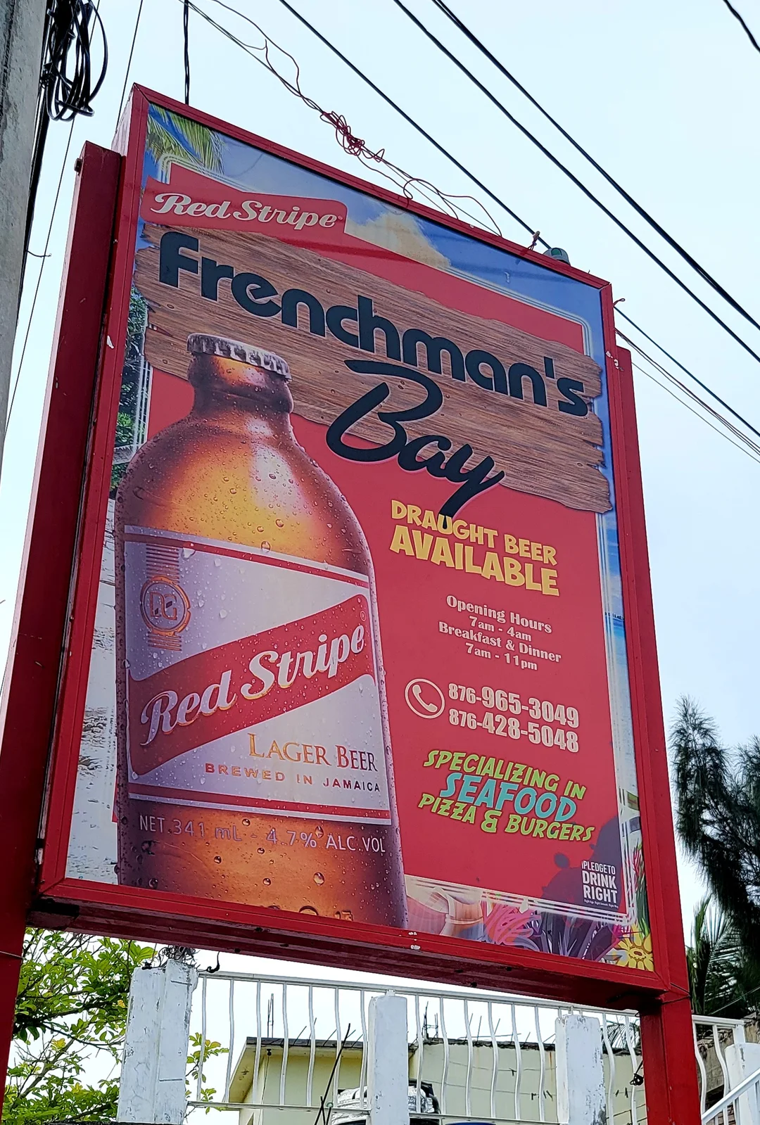 The sign for Frenchman’s Bay on a red frame: a large Red Stripe lager bottle beside the name on a wooden board, with the words Draught beer available, opening hours 7 am to 4 am, breakfast and dinner 7 am to 11 pm, the phone numbers 876 965 3049 and 876 428 5048, and Specializing in seafood, pizza and burgers