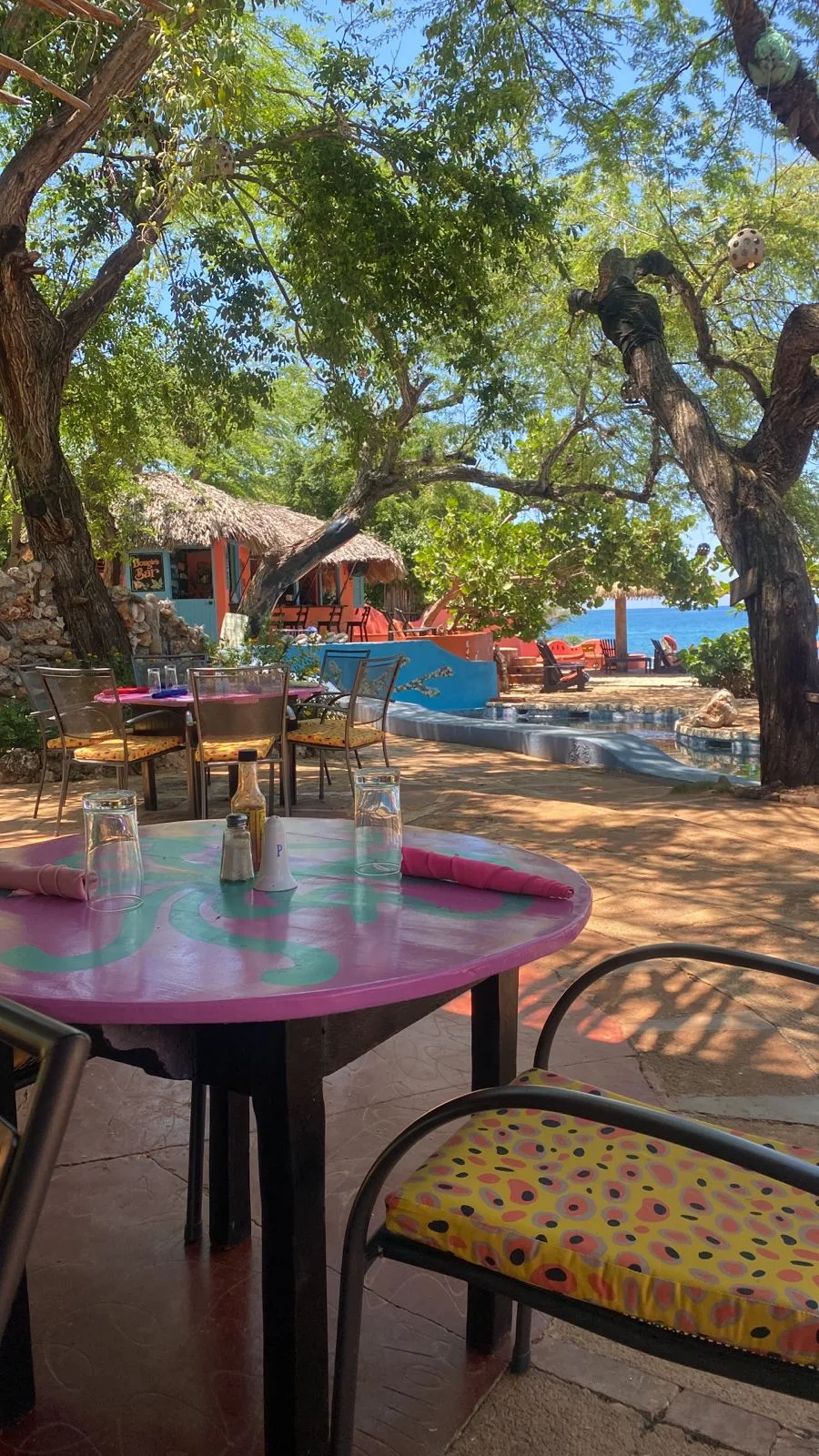Jakes Hotel Restaurant — A round purple and turquoise table set for a meal under shady trees, with Dougie’s Bar, the pool and the sea behind it