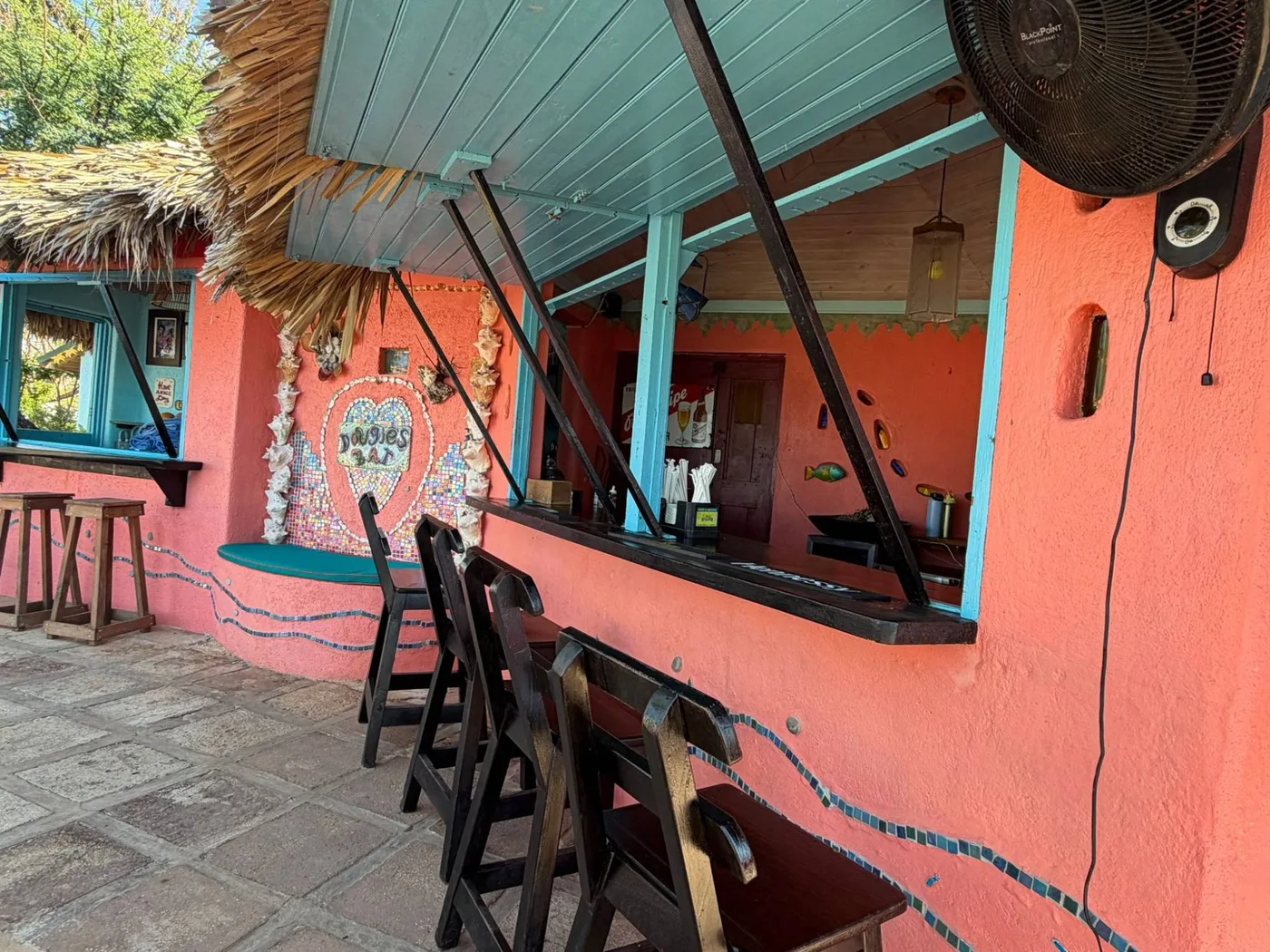 Dougie’s Bar at Jakes: a coral-pink bar with a thatched roof and a blue wooden shutter propped open, dark wooden bar stools and chairs, a heart of mosaic tiles and conch shells with the name Dougie’s Bar, and a fan on the wall
