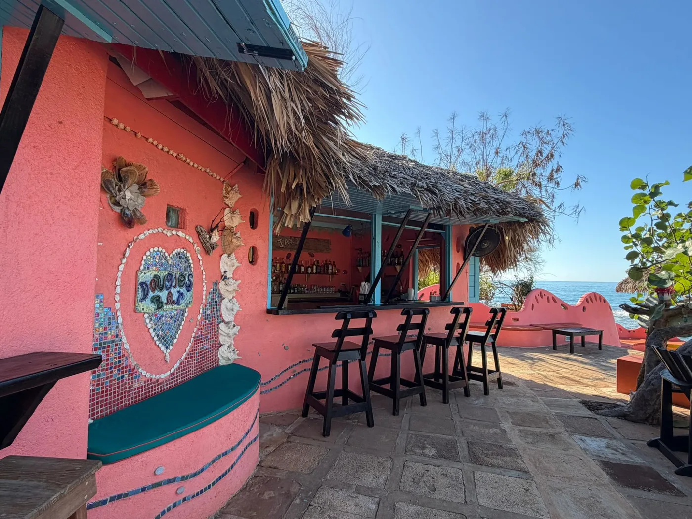 Dougie’s Bar beside the sea: bar stools along the coral-pink counter under the thatch, the mosaic heart and shell wall beside it, a curved pink wall around a terrace, and the blue sea beyond