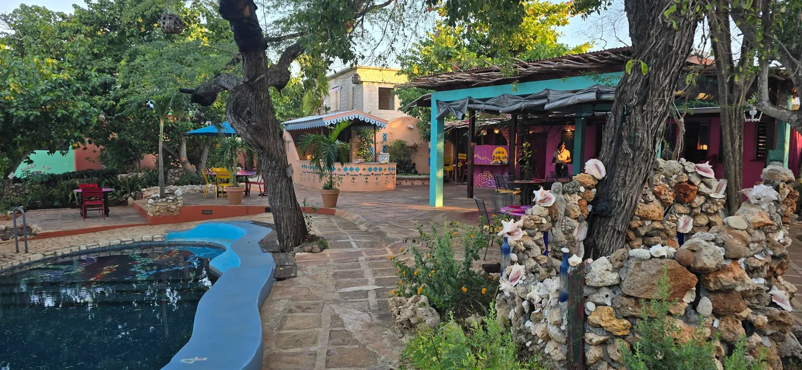 The pool and courtyard at Jakes Hotel: a curving blue-edged pool beside a flagstone path, shade trees, a stone wall studded with conch shells, and the restaurant’s turquoise and pink building with its tables and a waiter at work