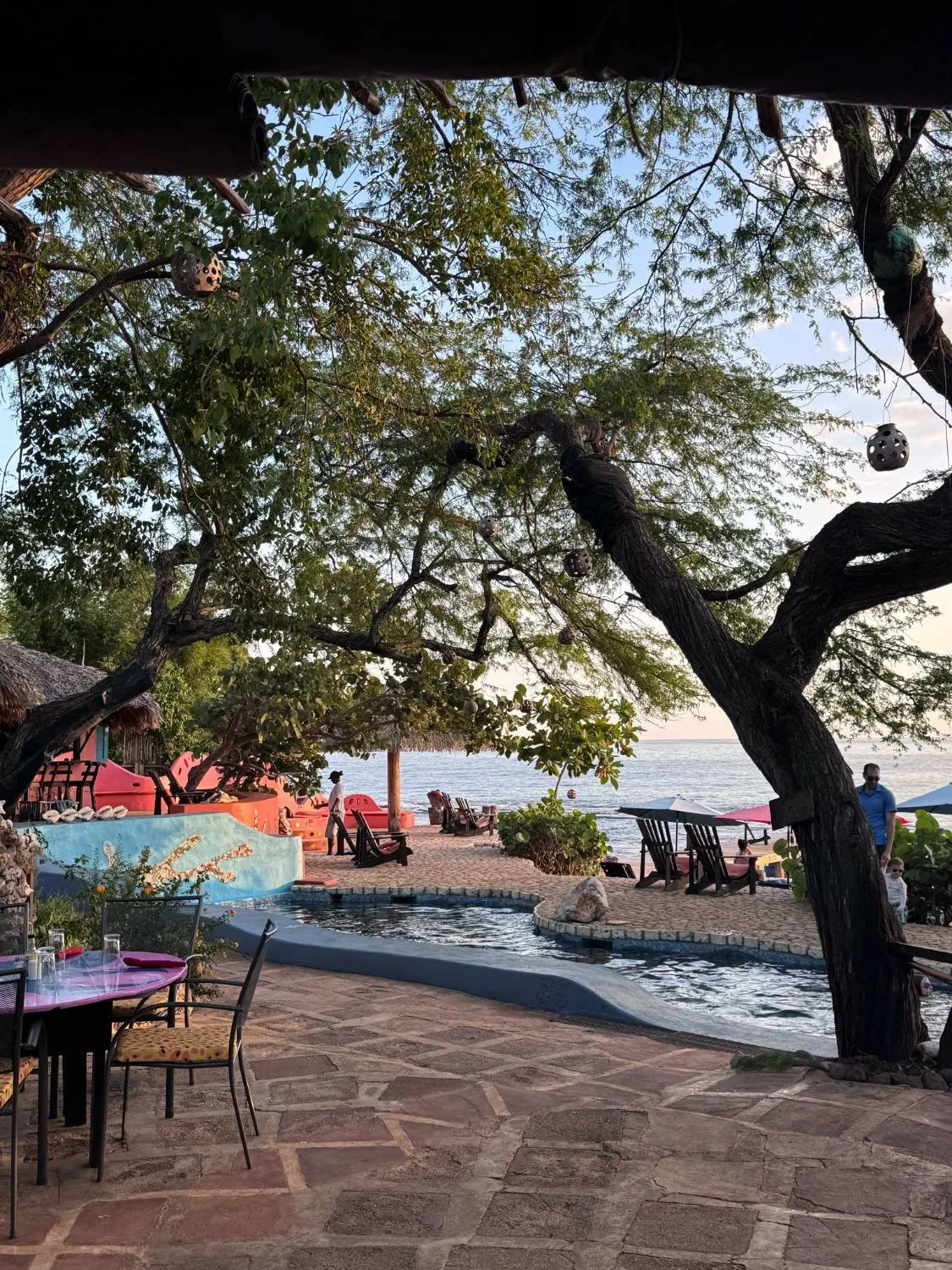 Jakes Hotel Restaurant — The pool and beach at dusk, seen from under the dining roof through the trees: the curved pool, sun chairs and umbrellas on the sand, the sea and a guest walking by