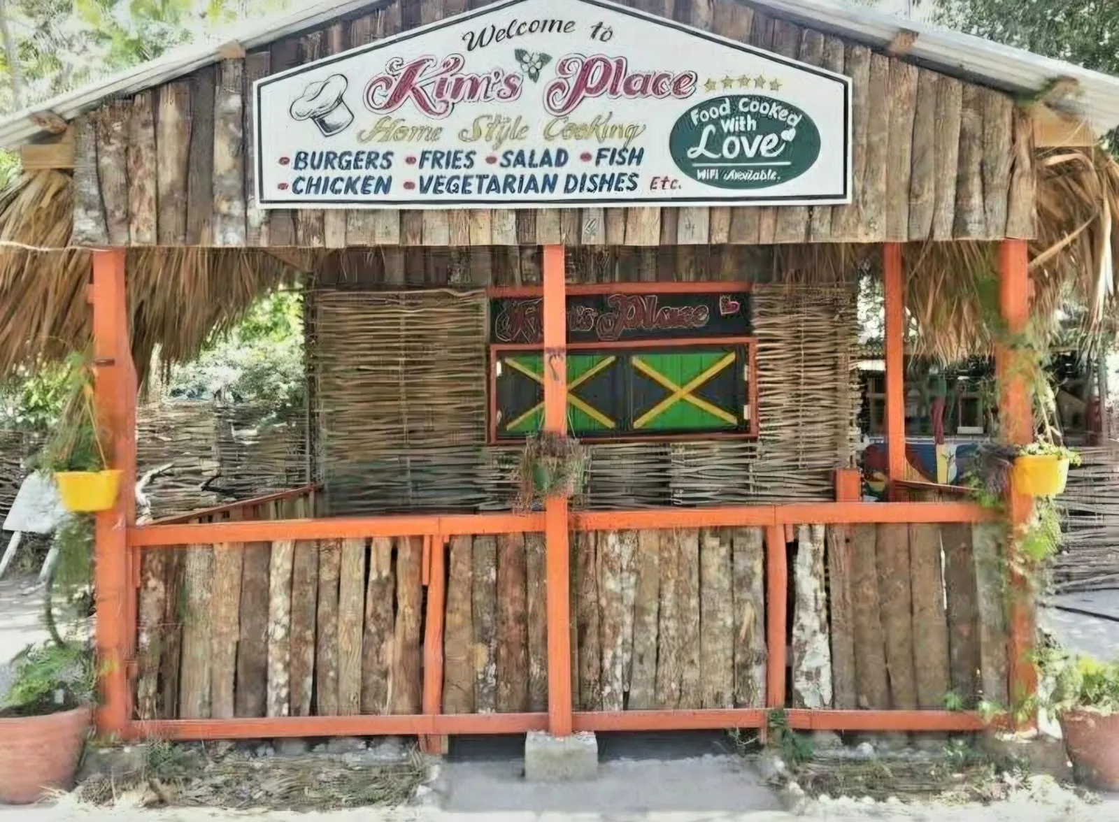 The front of Kim’s Place: a wooden cook shop with a thatched roof, orange posts and rails and a Jamaican flag on its shutters, under a sign reading Welcome to Kim’s Place, Home Style Cooking, Burgers, Fries, Salad, Fish, Chicken, Vegetarian Dishes etc., Food Cooked with Love, WiFi Available
