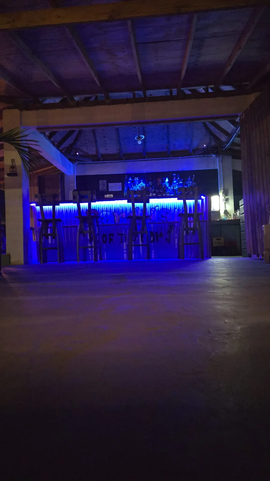 Pardy’s Rooftop Bar — The bar seen across the rooftop floor, lit in blue and purple, with four stools at the counter