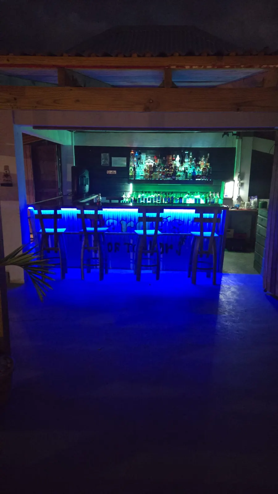Pardy’s Rooftop Bar — The bar counter lit in blue, with four wooden stools and two shelves of bottles behind it