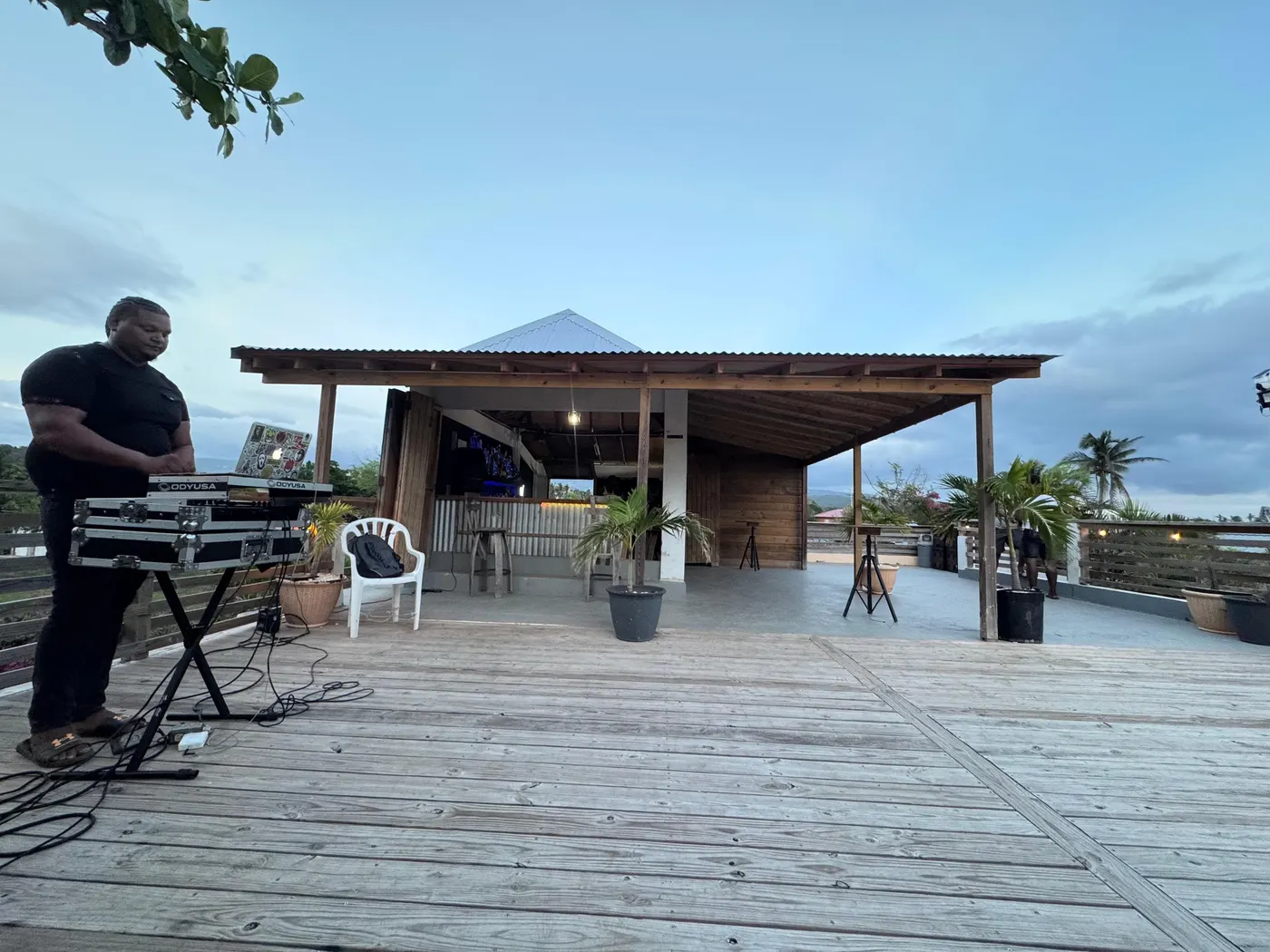 A DJ at his decks on the wooden deck at dusk, with the open-sided bar, its corrugated roof and the rooftop behind him