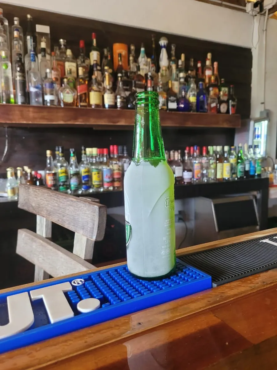 Pardy’s Rooftop Bar — An ice-cold green Heineken bottle, frosted over, on a blue bar mat, with shelves of spirits behind the bar