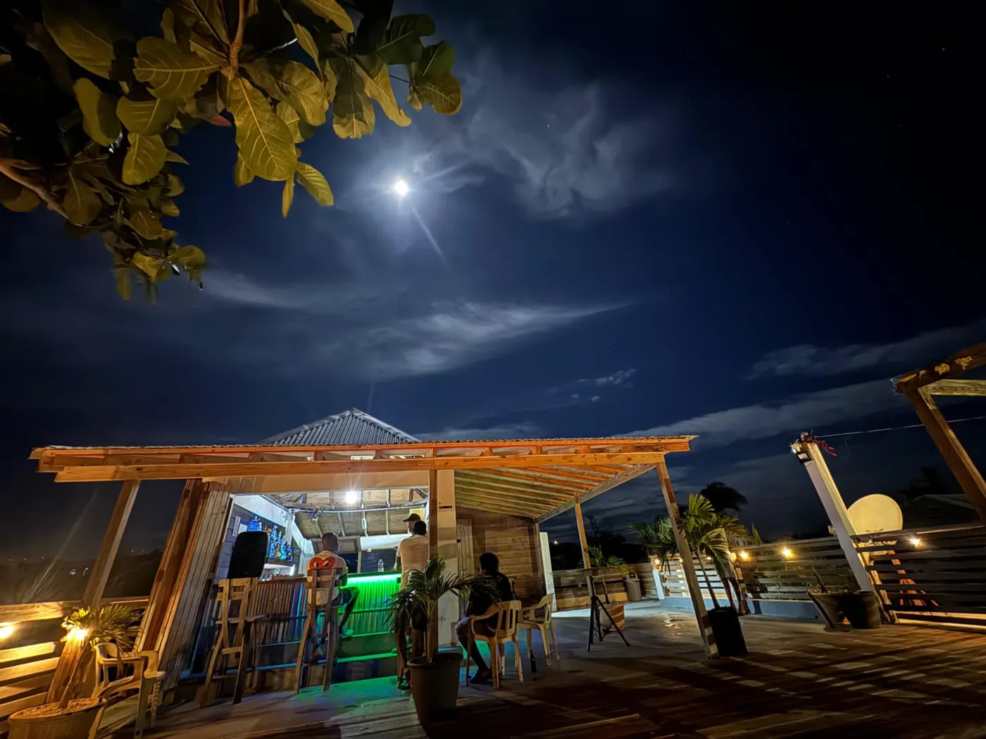 Pardy’s Rooftop Bar — Pardy’s Rooftop Bar at night: an open-sided wooden bar under a corrugated roof, lit green under the counter, with guests on plastic chairs, a full moon above and a sea-almond branch in the corner
