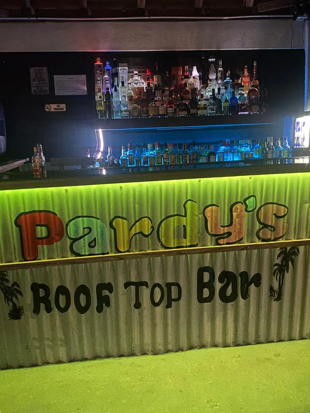 The front of the bar, painted on corrugated metal: Pardy’s in red, blue, yellow and orange letters and Roof Top Bar beneath, between two palm trees, lit green, with shelves of bottles behind
