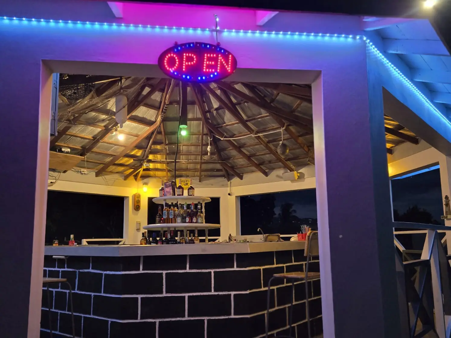 The bar at the Treasure Beach Inn at night: a red and blue OPEN sign over the doorway, blue and pink lights along the roof, bottles on a tiered stand behind a dark brick counter, and tall stools
