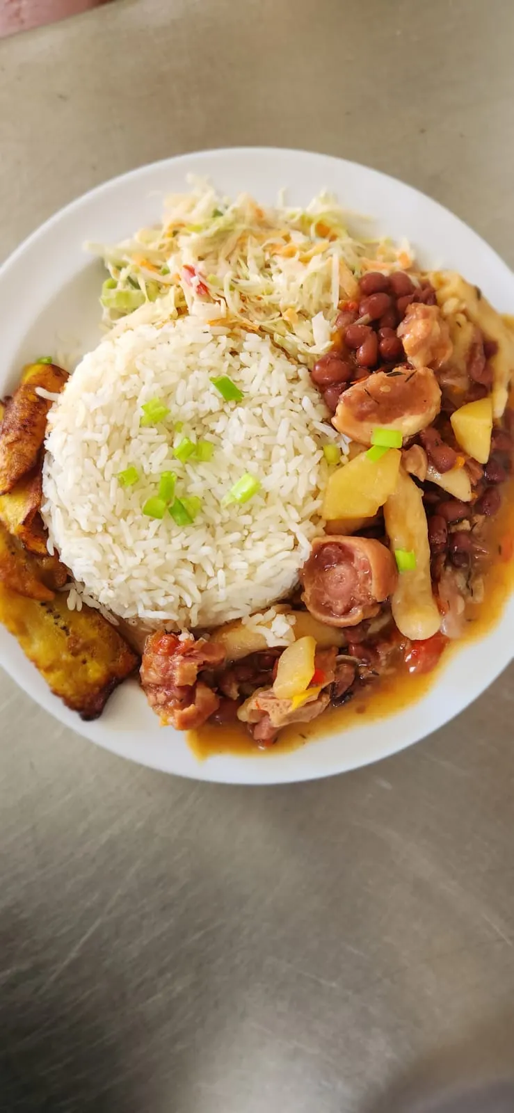 A plate of stew peas with white rice topped with scallion, fried ripe plantain and a cabbage and carrot slaw