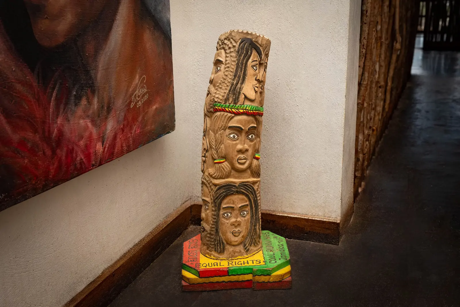 A carved wooden totem of three faces on a red, gold and green base, standing in a corridor beside a painting