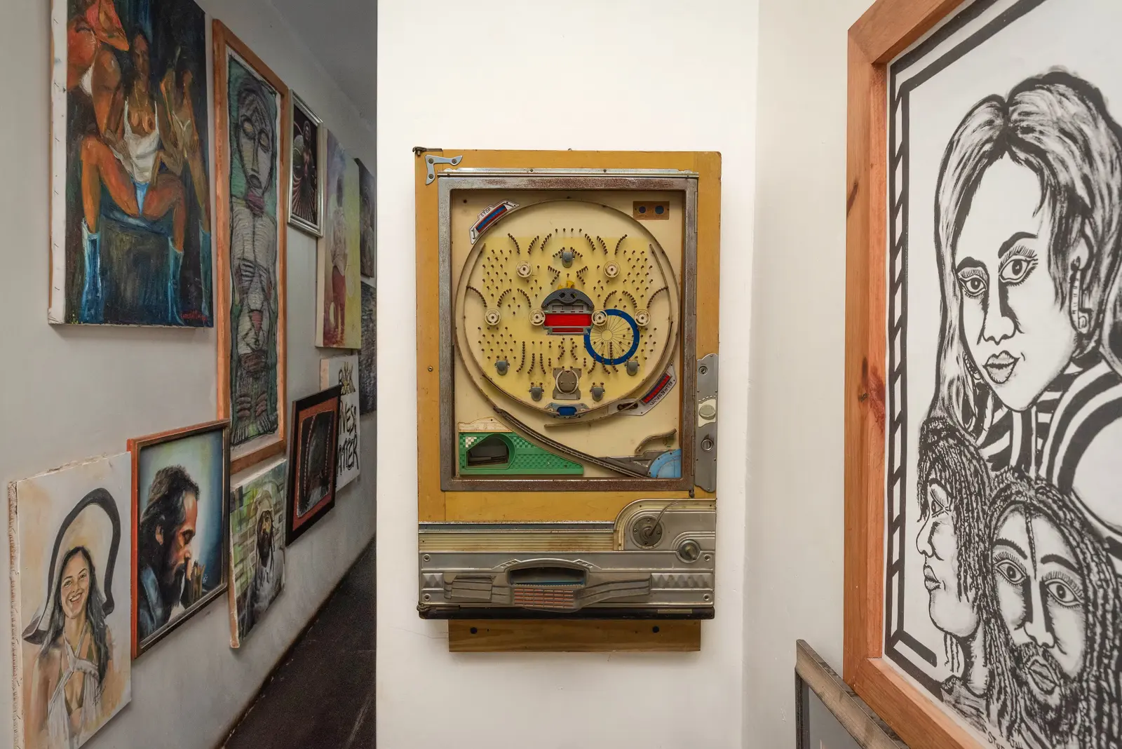 A wall of paintings and drawings along a passage, with a vintage pinball-style machine and a large black-and-white portrait drawing