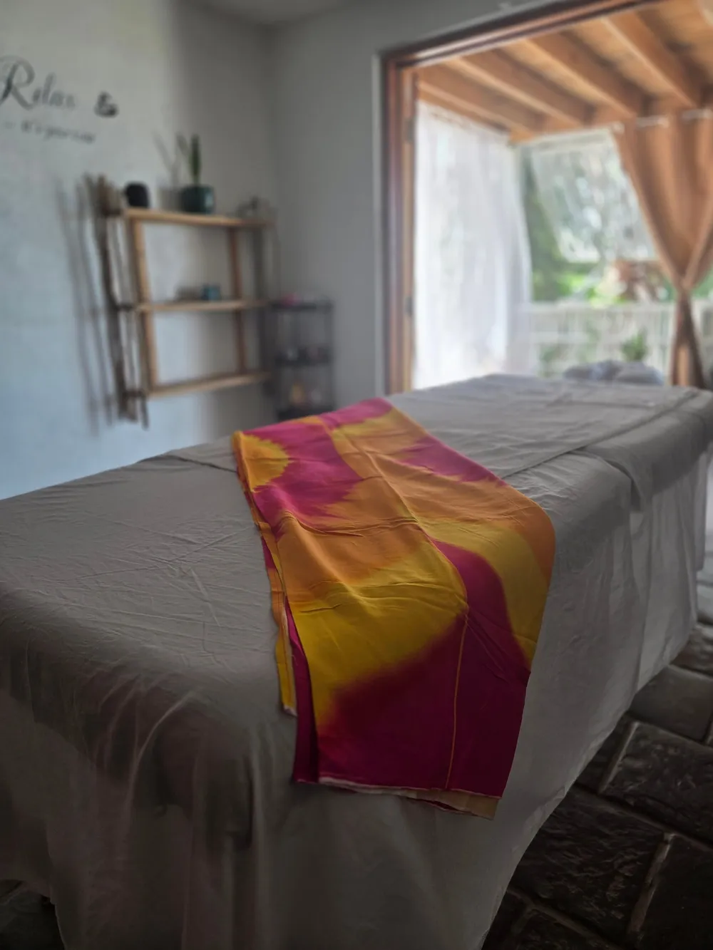 A massage table made up with grey sheets and a bright pink, orange and yellow cloth, beside an open door to a shaded veranda with curtains and plants