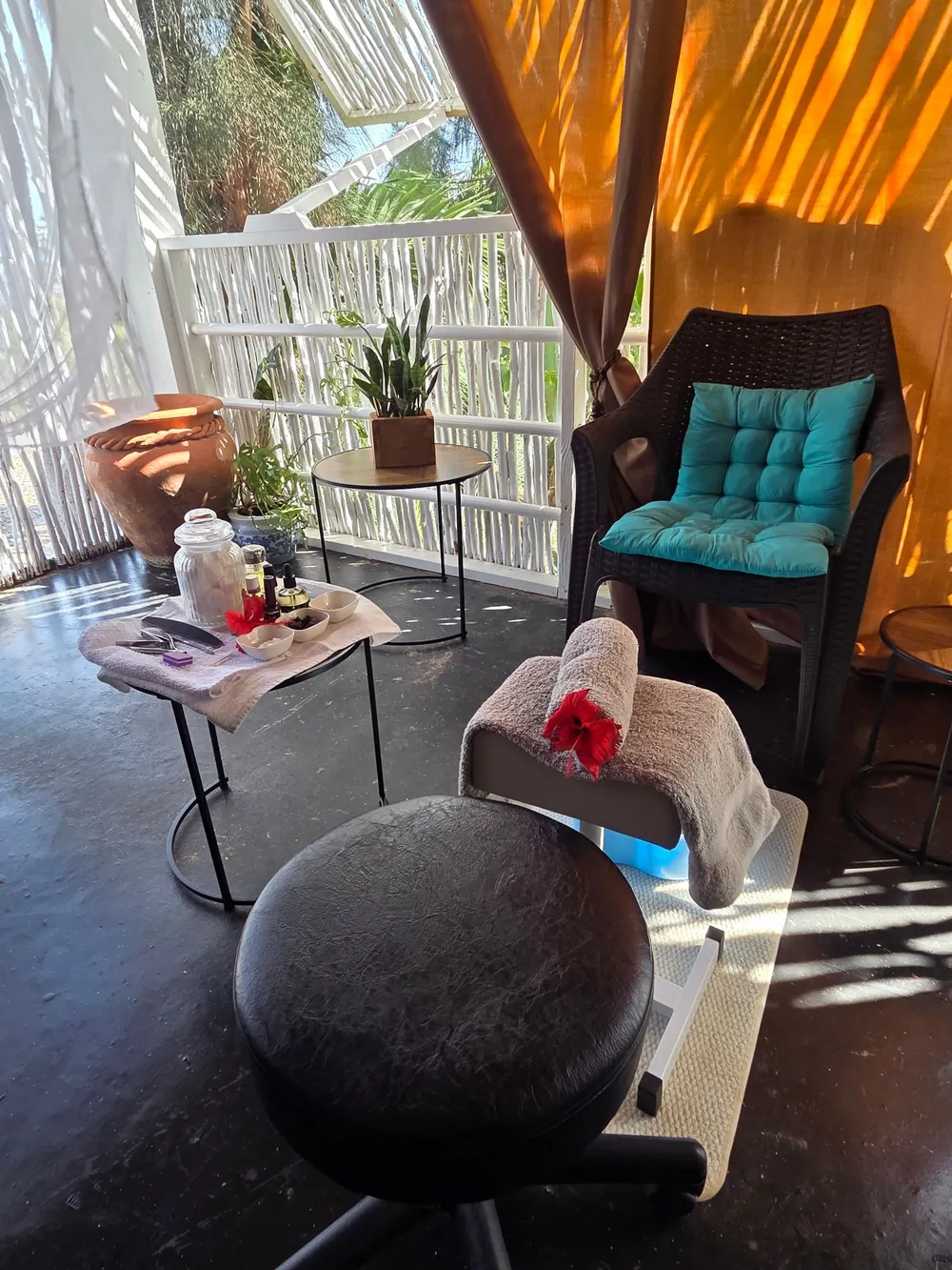 A pedicure corner on a shaded veranda: a footrest with a rolled towel and a red hibiscus flower, a stool, a small table of nail tools, and a chair with a turquoise cushion, with palm trees beyond the white railing
