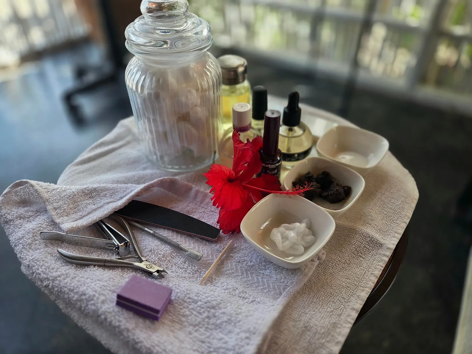 A tray of manicure tools on a white towel: nail files, clippers and cuticle tools, small dishes of cream and scrub, oils and polish, a jar of cotton pads and a red hibiscus flower