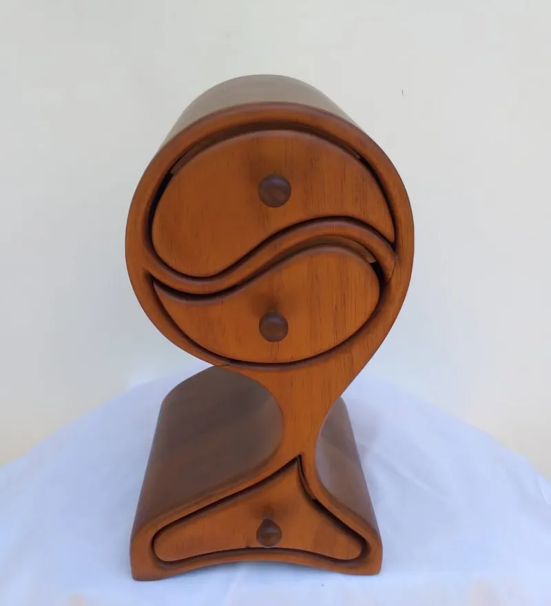 A tall wooden jewellery box whose two upper drawers make a yin yang circle, above a third drawer in a wave-shaped base