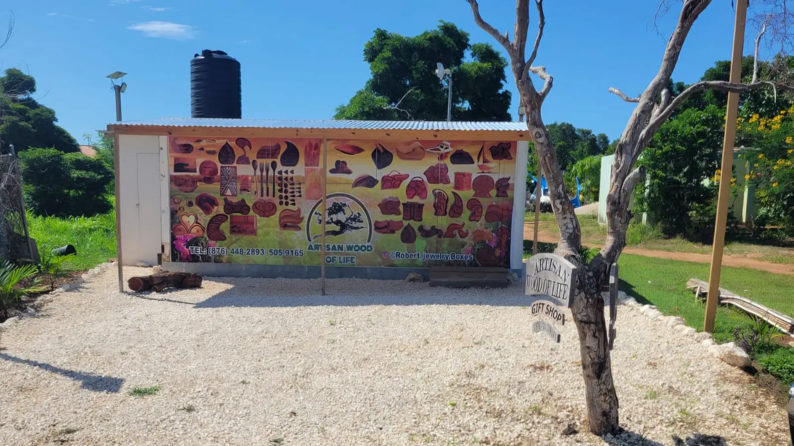The Artisan Wood of Life shop head on: a small white building with a tin roof, its front wall covered in a printed picture of the carved boxes and wooden goods for sale, with the shop's name, a phone number and an Instagram handle, and a tree beside a gravel yard in the sunshine