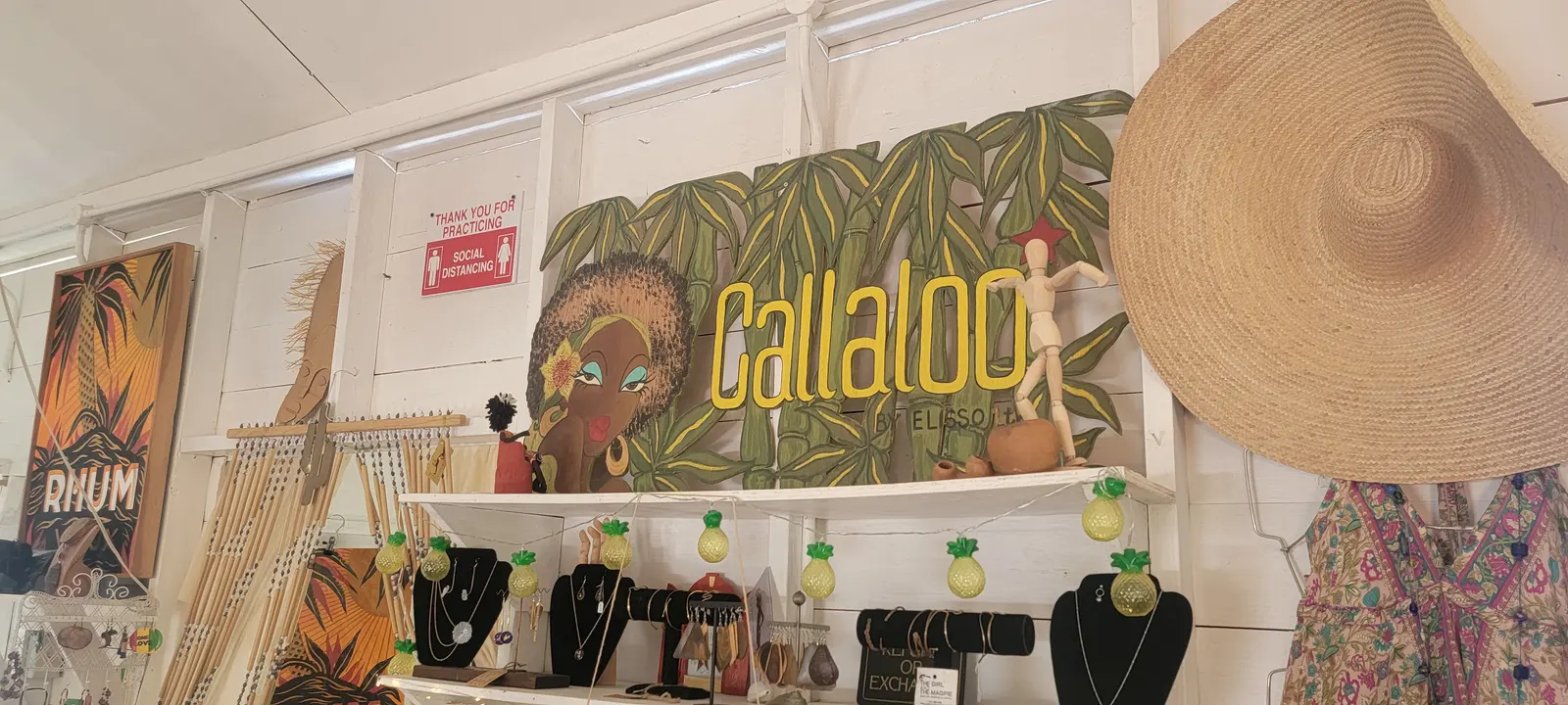 Inside the shop: a painted Callaloo sign above shelves of jewellery on black stands, a straw hat, framed prints and a rack of dresses