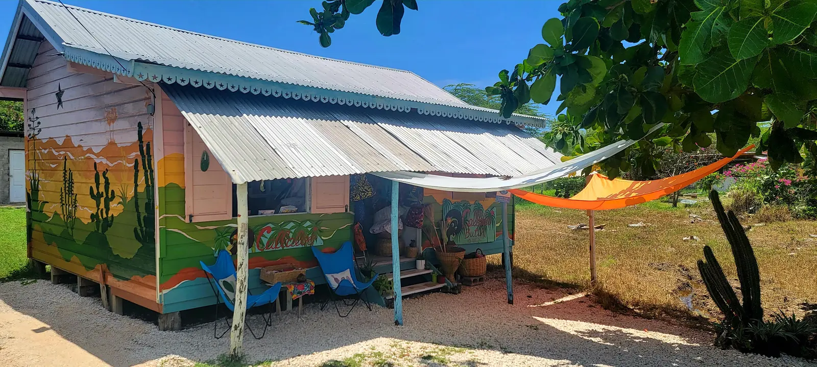 The Callaloo Butik shop, a small wooden building with a mural of cacti and hills in green, yellow and orange, a blue-trimmed porch with two butterfly chairs, baskets and an orange sunshade