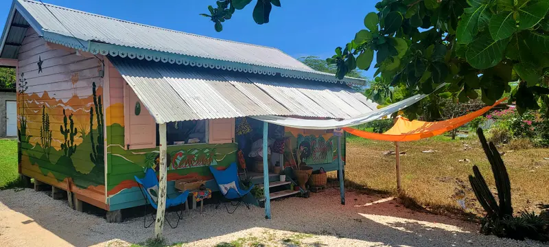 The Callaloo Butik shop, a small wooden building with a mural of cacti and hills in green, yellow and orange, a blue-trimmed porch with two butterfly chairs, baskets and an orange sunshade