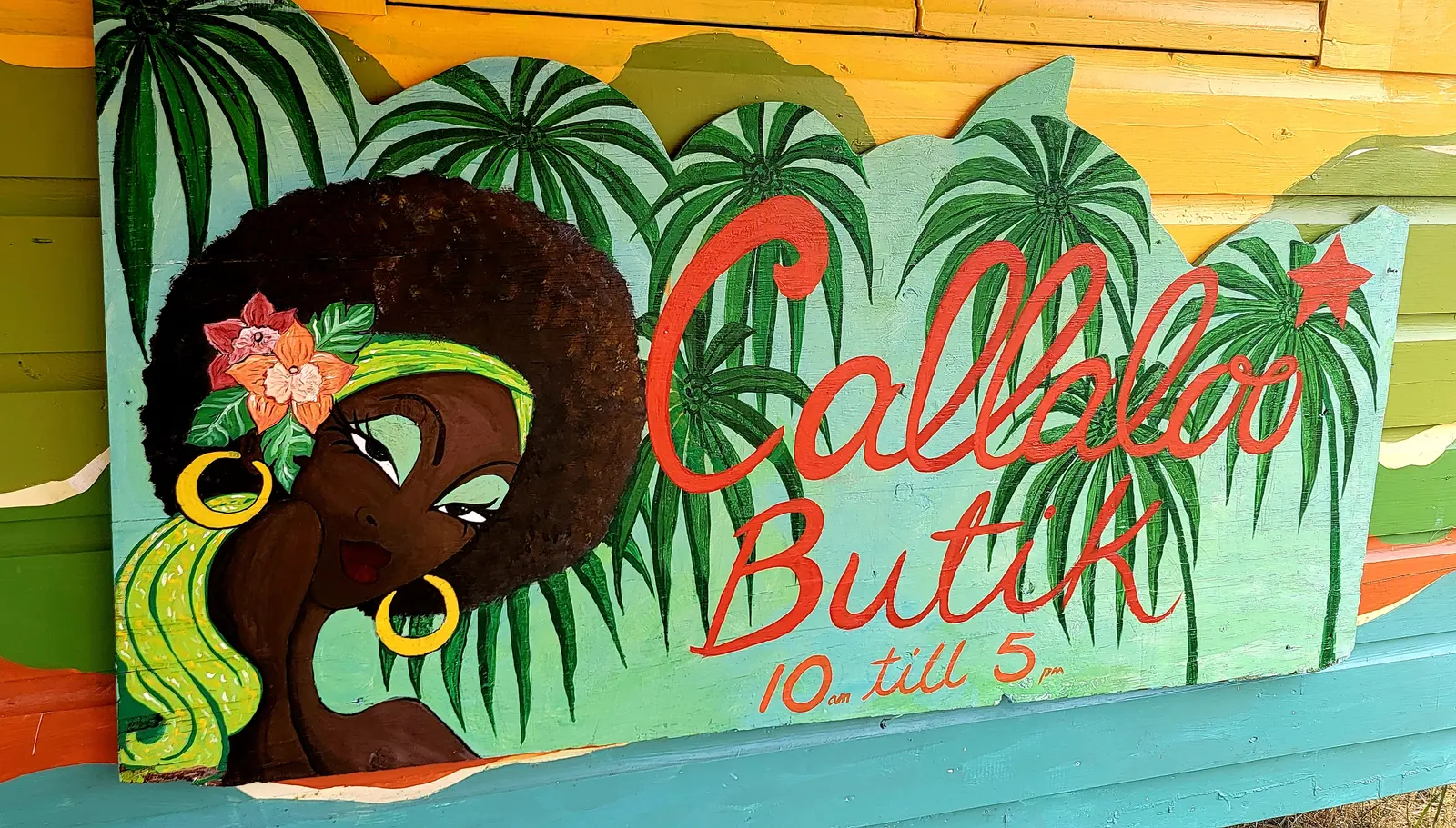 The painted Callaloo Butik sign: a woman with an afro, a green headband, gold hoop earrings and an orange flower, with the name in red script among palm leaves, and the hours 10 am till 5 pm