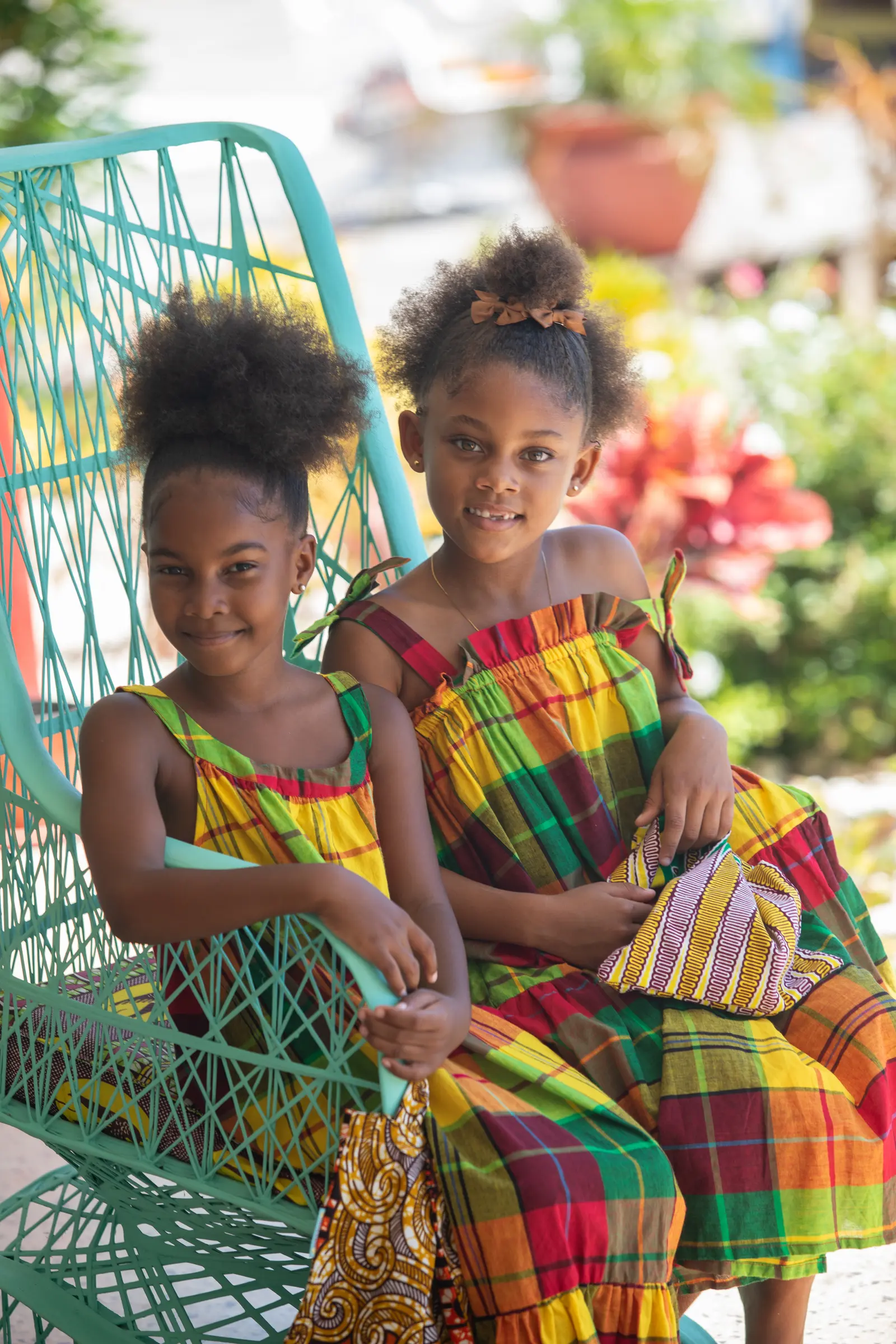 Two girls sitting in a turquoise woven chair, in madras-check dresses in yellow, green and red, one holding a pouch in yellow African-print cloth