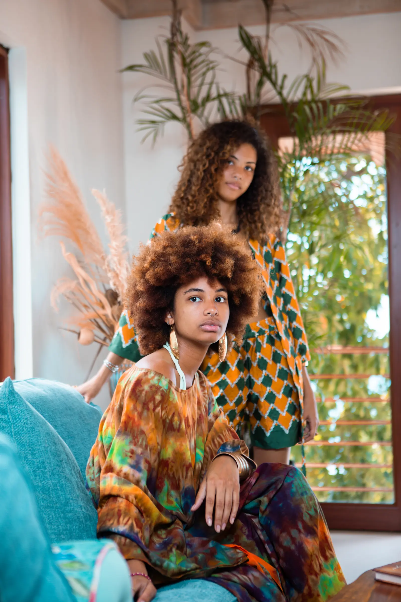 Two young women among palms and plants, one seated on a teal sofa in an orange, green and purple tie-dye top and trousers, the other standing behind in an orange and green diamond-print jacket and shorts