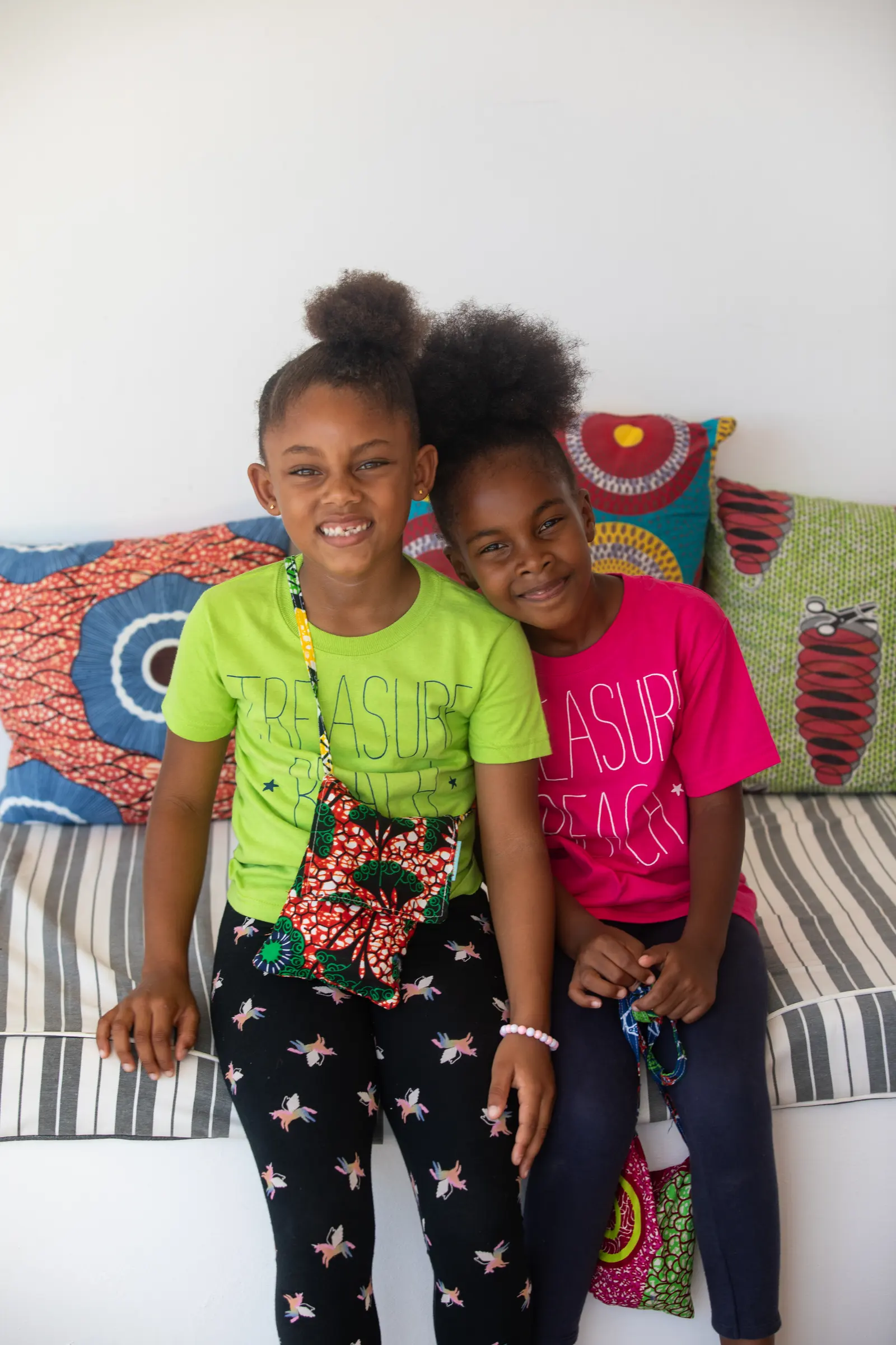 Two girls in Treasure Beach T-shirts, one green and one pink, with small African-print bags, on a sofa with African-print cushions