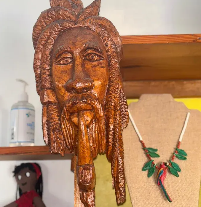 A carved wooden head with long locks and a beard, beside a necklace with a painted wooden parrot on a linen stand, and a cloth doll on the shelf below