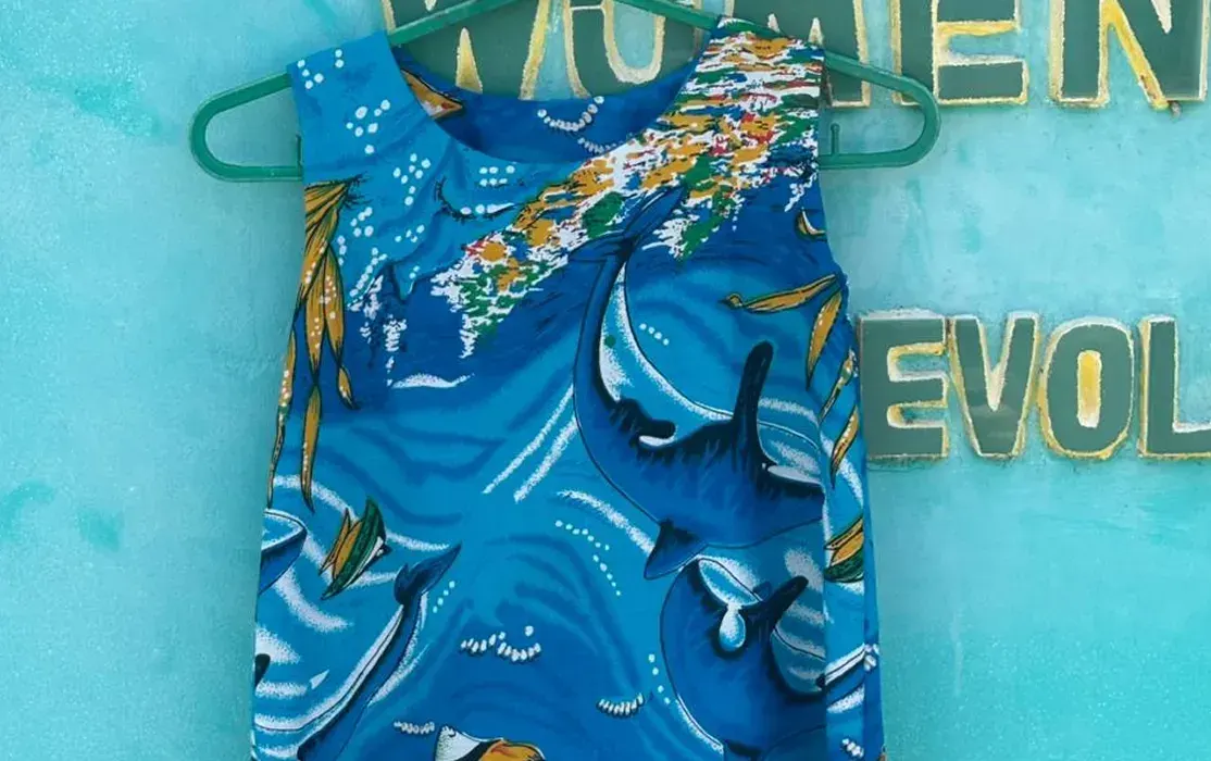 A child's sleeveless dress in a blue print of dolphins and fish, hung against a turquoise wall