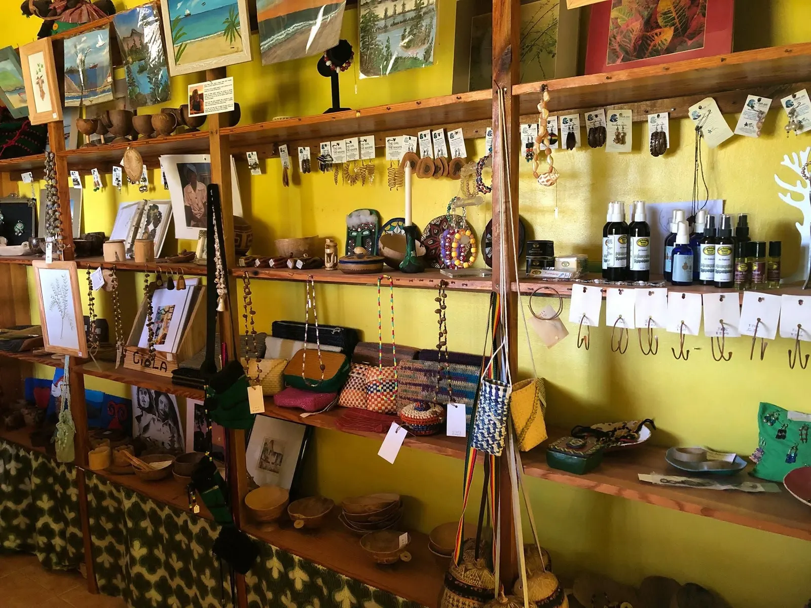 Shelves in the shop against a yellow wall: earrings and necklaces on cards, calabash bowls and cups, woven purses and baskets, paintings and prints, and bottles of natural sprays and oils