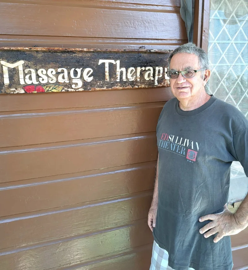 Joshua Lee Stein standing beside a carved wooden Massage Therapy sign on a brown wooden wall