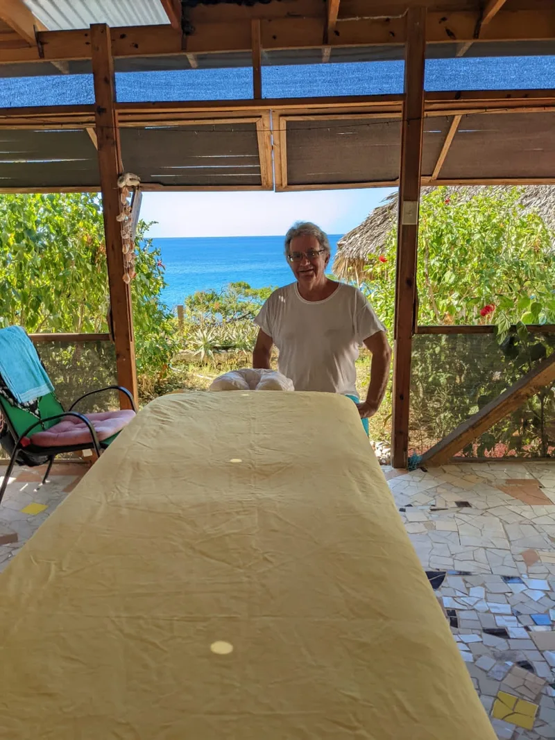 Joshua Lee Stein behind a massage table in an open-sided wooden room, with the blue sea and green garden behind him