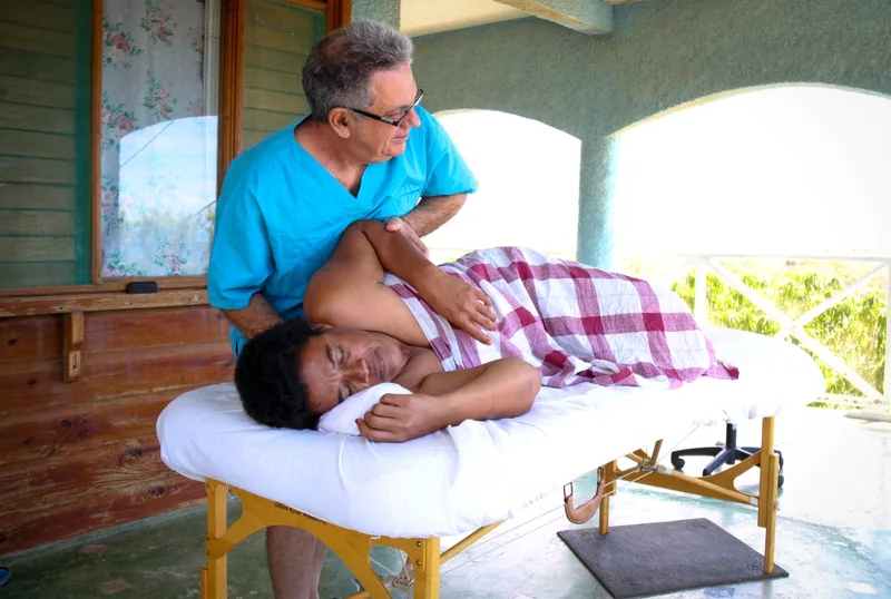 Joshua Lee Stein treating a woman lying on her side on a massage table on a shaded veranda, steadying her shoulder