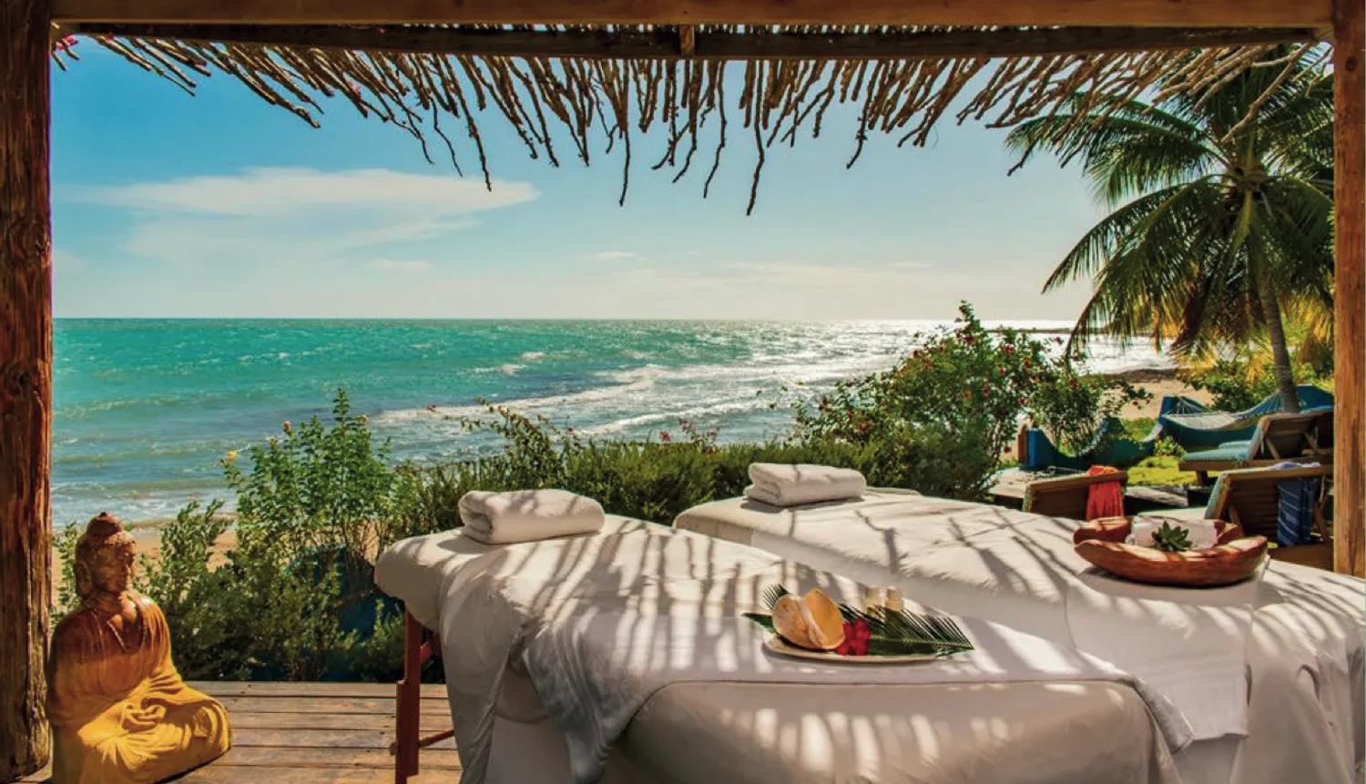 A massage table with rolled towels under a thatched open-sided cabana, looking out over the turquoise sea and a palm, with a golden Buddha statue on the deck