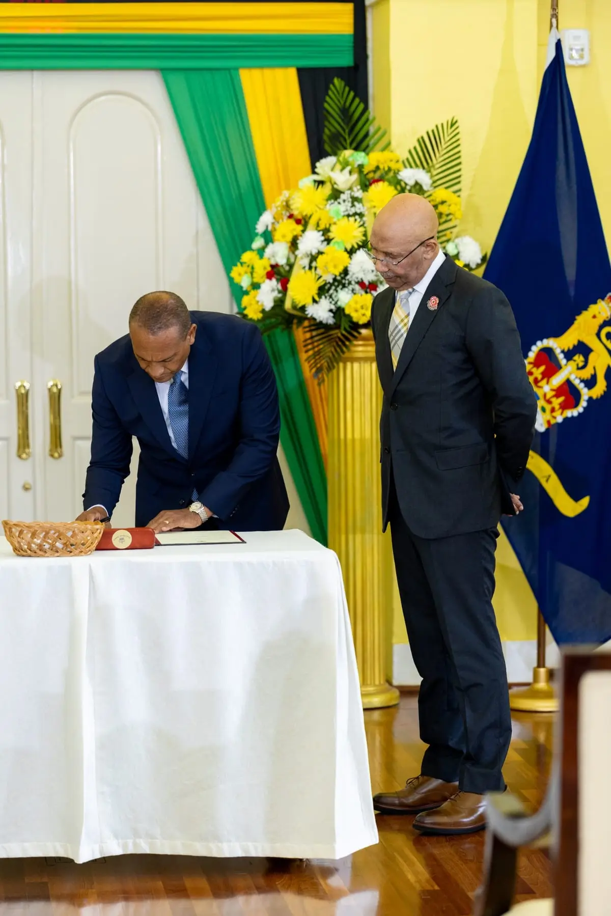 Bernard Henry, on the left, bends over a table with a white cloth to sign his documents while Governor General Sir Patrick Allen, on the right, looks on with his hands behind his back